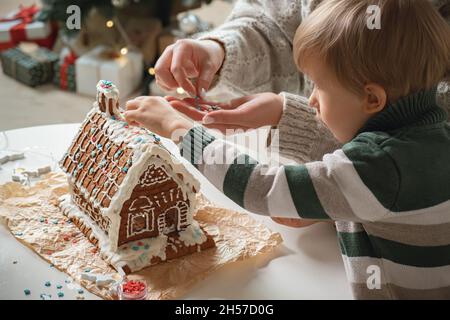 Ragazzino con madre che decora insieme casa di pan di zenzero di natale, attività di famiglia e tradizioni a Natale e Capodanno Foto Stock