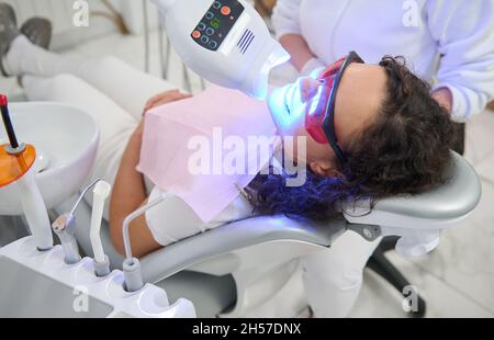 Attraente giovane paziente donna seduta in sedia dentista, con bocca aperta che riceve il trattamento di sbianca con moderna lampada a luce ultravioletta speciale per Foto Stock