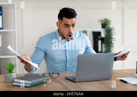 Scioccato uomo d'affari arabo con documenti che guardano lo schermo del laptop, urlando WTF, facendo errori nel progetto in ufficio Foto Stock