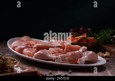 Pollo crudo biriyani tagliato senza pelle disposto su stoviglie bianche con ingredienti posti sullo sfondo con fondo rustico in legno. Foto Stock