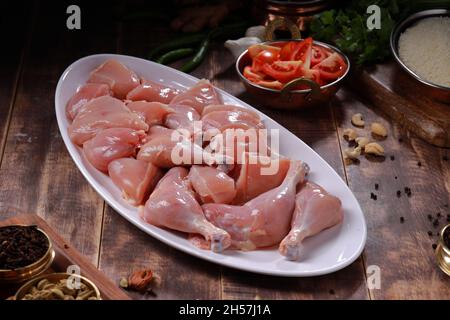 Pollo crudo biriyani tagliato senza pelle disposto su stoviglie bianche con ingredienti posti sullo sfondo con fondo rustico in legno. Foto Stock