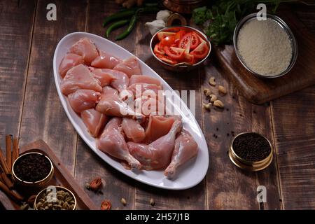 Pollo crudo biriyani tagliato senza pelle disposto su stoviglie bianche con ingredienti posti sullo sfondo con fondo rustico in legno. Foto Stock