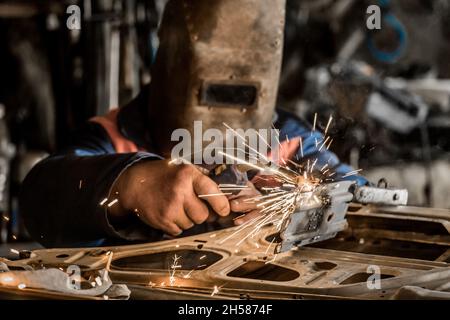 Il saldatore in una maschera di protezione è impegnato in lavori di saldatura della, scintilla e riparazione della porta dell'auto nel negozio dell'impianto industriale. Foto Stock