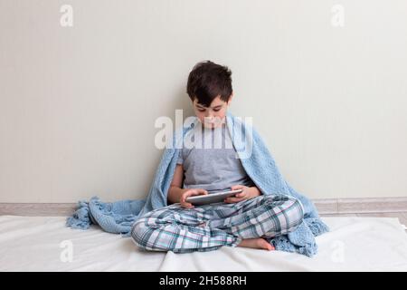 Un ragazzo di bruna, un adolescente in pigiama grigia, sul pavimento, sorride, usa un tablet digitale Foto Stock