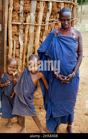 Africa, Tanzania, 1976. Ujamaa villaggio Maasai persone ancora vivono nelle loro case tradizionali. Maasai donna e due dei suoi figli in piedi di fronte alla loro capanna. Foto Stock