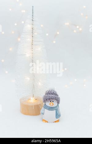 Figurine decorative a tema natalizio. La statuetta di un pinguino in un cappello a maglia su sfondo bianco. Decorazioni festive, calde luci bokeh Foto Stock