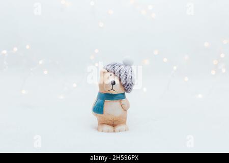 Figurine decorative a tema natalizio. La statuetta di un orso in un cappello a maglia su sfondo bianco. Decorazioni festive, calde luci bokeh Foto Stock