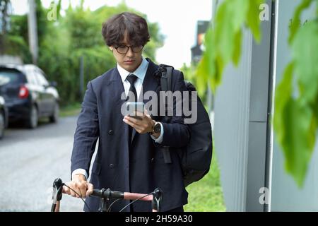 Giovane uomo d'affari asiatico che guarda i messaggi sul cellulare mentre si guida in bicicletta in città. Foto Stock