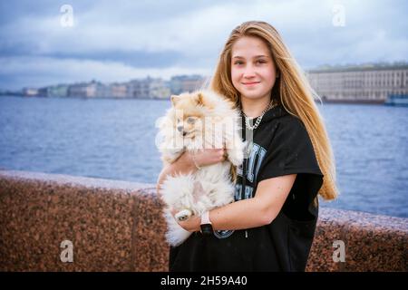 Ragazza allegra e felice adolescente tiene e abbraccia Pomeranian spitz sul terrapieno in città il giorno nuvoloso, fuoco selettivo. Giovane donna caucasica con il suo amato animale domestico sulla riva del fiume nel vento sorridendo Foto Stock