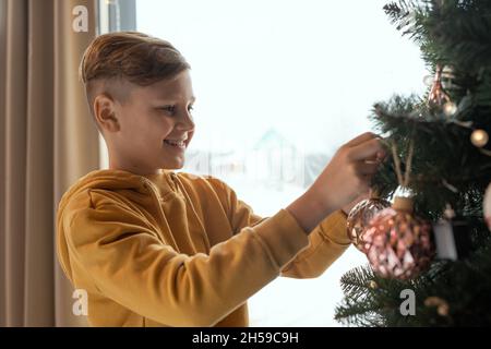 Ragazzo felice in previsione di Capodanno in piedi all'albero di Natale e giocattolo appeso sul ramoscello Foto Stock