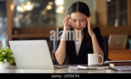 Business manager femminile asiatica con mal di testa. Lavorare alla scrivania di fronte al notebook che soffre e affrontare il problema del lavoro. Foto Stock