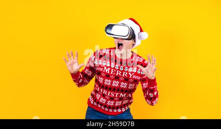 Giovane uomo latino giocatore in realtà virtuale cuffia gadget indossando Santa cappello e maglione di Natale su sfondo giallo. Natale e inverno vr technolo Foto Stock