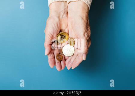 Le mani di femakes rugose tengono una manciata di bitcoins e monete d'oro. Sfondo blu. Disposizione piatta. Il concetto di banca e criptovaluta. Foto Stock