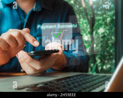 Pagamento riuscito, parole e carrello della spesa e segno di spunta sullo schermo virtuale dello smartphone vicino al computer portatile. Uomo che usa il telefono cellulare, on Foto Stock