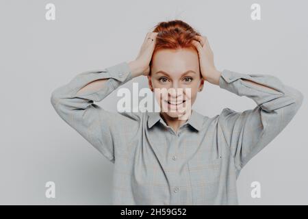 Emozionata donna dai capelli rossi che tiene la testa nelle mani e guarda la fotocamera con un'espressione sorpresa Foto Stock