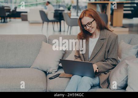 Donna in occhiali seduti su un comodo divano nella sala d'ufficio mentre chiacchiera sul computer portatile Foto Stock