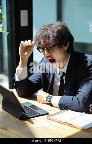 Il salarista stanco lavora sul suo tablet digitale e si sente assonnato alla sua workstation Foto Stock