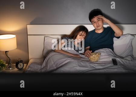 giovane che guarda la televisione nel letto con la moglie, espressione addormentata e stanca che soffre di insonnia Foto Stock