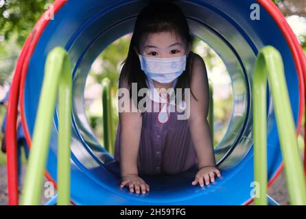 Una giovane ragazza asiatica carina, che indossa una maschera bianca, gioca da sola all'aperto, strisciando attraverso un tunnel colorato in una palestra nella giungla di un parco giochi in un parco Foto Stock