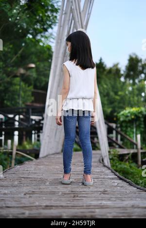 La vista posteriore di una giovane ragazza asiatica carina in camicia bianca e jeans blu che camminano lungo un lungo percorso di legno con piantagione verde lungo il lato. Foto Stock