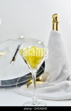 Un bicchiere festivo con uva sullo sfondo di un orologio sfocato e una bottiglia di champagne. La tradizione spagnola di mangiare dodici uve per celebrare il nuovo Foto Stock