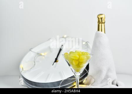 Bicchiere con uva sullo sfondo di un orologio sfocato e una bottiglia di champagne. La tradizione spagnola di mangiare dodici uve per festeggiare l'anno nuovo. Foto Stock