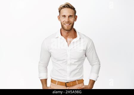 Bel macho uomo con capelli biondi e barba, sorridente e guardando sicuro alla macchina fotografica, indossando una camicia bianca, in piedi sullo sfondo dello studio Foto Stock