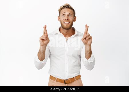 Il giovane uomo fiducioso e nervoso incrocia le dita, pregando o facendo il desiderio, guardando in alto con la speranza, in piedi su sfondo bianco Foto Stock