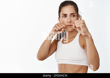 Sportivo forte e concentrato in piedi con pugni, pugni di pugilato di allenamento, combattente femminile facendo esercizi, in piedi su sfondo bianco Foto Stock