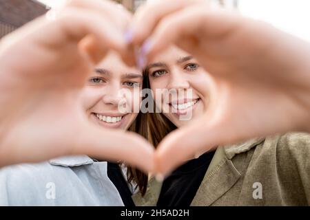 Volti di due sorelle gemelle felici che ti guardano con sorrisi toothy attraverso il cuore della mano mentre ti trovi di fronte alla fotocamera Foto Stock