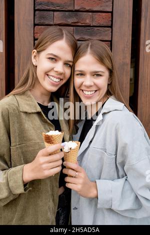 Belle sorelle gemelle con lunghi capelli biondi in piedi contro la parete di un edificio moderno e avere il icecream di fronte alla macchina fotografica Foto Stock