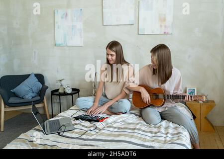 Due ragazze adolescenti felici che registrano la musica a casa mentre una di loro suona la chitarra e l'altra premendo i tasti della tastiera del piano Foto Stock