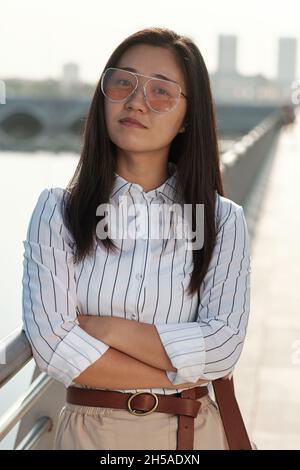 Giovane donna d'affari fiduciosa in occhiali, maglietta a righe e gonna beige che mantiene le braccia incrociate dal petto Foto Stock