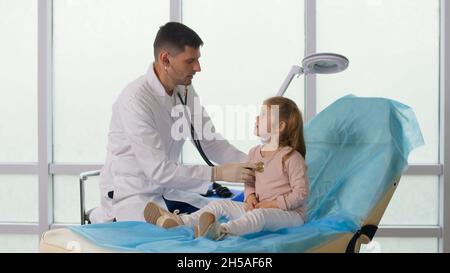 Il paziente figlio visita l'ufficio medico. Il medico ascolta i polmoni del bambino con uno stetoscopio, parla con la bambina. Foto Stock