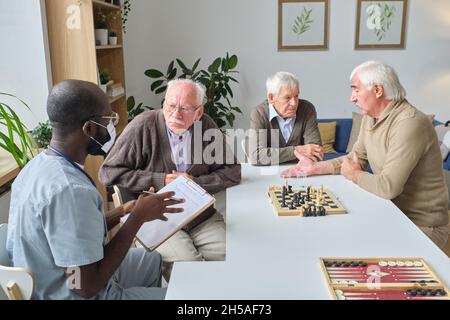 Medico di sesso maschile che lavora in casa di cura, egli prescrivendo medicina a uomo anziano mentre si siedono al tavolo con altri uomini che giocano a scacchi in background Foto Stock