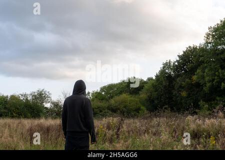 Un'ambientazione suggestiva di un personaggio con cappuccio, torna alla fotocamera. In piedi in un campo che guarda verso il cielo. Foto Stock