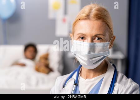 Primo piano di una donna matura medico in maschera protettiva guardando la macchina fotografica mentre si tratta il bambino malato in ospedale Foto Stock