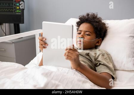 Ragazzo malato sdraiato a letto e leggere online libro su tablet digitale durante il trattamento in ospedale Foto Stock