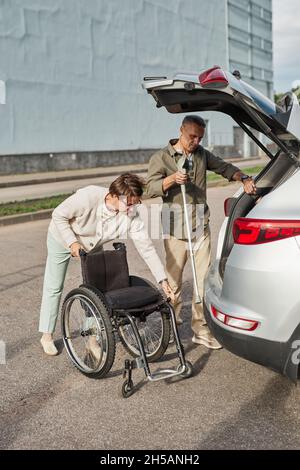 Ritratto a tutta lunghezza di coppia matura caricando la sedia a rotelle al bagagliaio dell'automobile nel parcheggio all'aperto Foto Stock