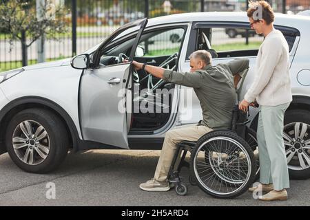 Ritratto di donna aiutare l'uomo in sedia a rotelle entrare in auto nel parcheggio all'aperto, spazio copia Foto Stock