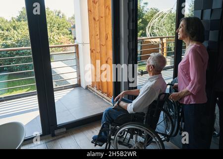 Uomo con disabilità in sedia a rotelle che guarda il cortile dalla finestra Foto Stock