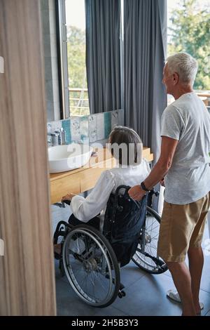 Età maschile che tiene la sedia a rotelle di disabili da dietro Foto Stock