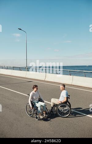 Ritratto verticale di coppia adulta in sedia a rotelle chiacchierando all'aperto e godendo la vita della città, spazio copia Foto Stock