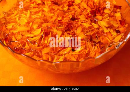 Primo piano di petali di fiori di Marigold in una ciotola di vetro trasparente riempita d'acqua su sfondo arancione Foto Stock