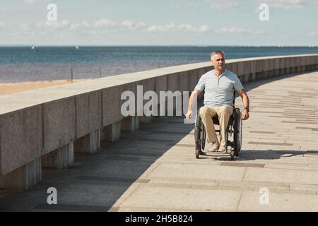 Ritratto a tutta lunghezza di uomo adulto in sedia a rotelle godendo all'aperto sul lungofiume, spazio copia Foto Stock