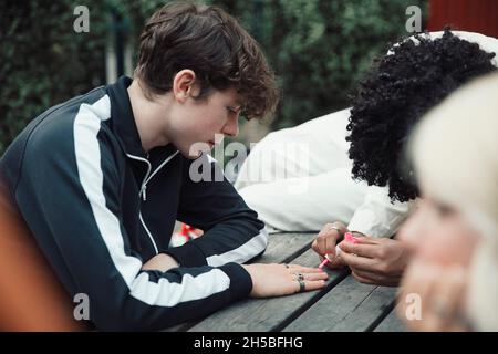 Ragazzo adolescente che applica smalto rosa per unghie a un amico maschio a tavola Foto Stock