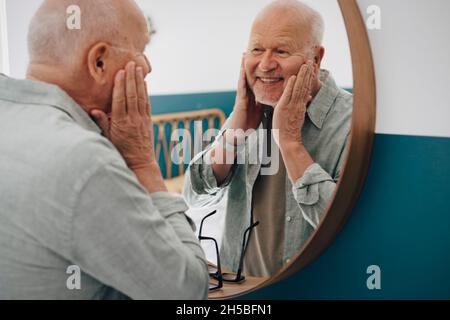 Uomo anziano sorridente che tocca la pelle mentre guarda nello specchio Foto Stock