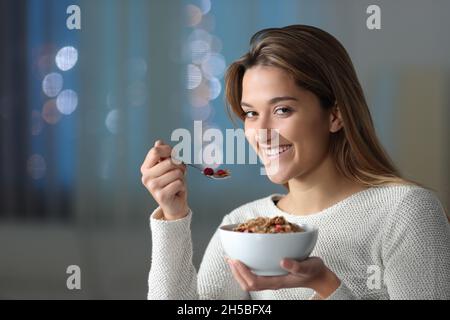 Donna felice che guarda la macchina fotografica che mangia cereali la notte a casa Foto Stock