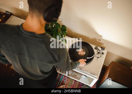 Il giovane uomo millenario della corsa mista mette Needle sul disco in vinile, ascoltando la musica con le cuffie sul suo appartamento. Vista aerea delascolto di musica Foto Stock