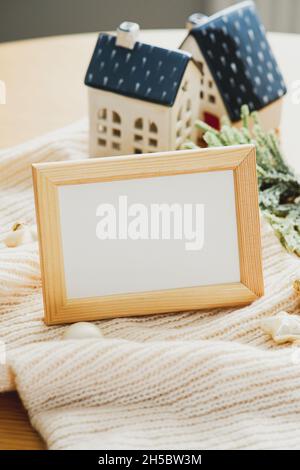 Hygge, concetto estetico casa accogliente. Cornice fotografica mockup, baubles beige Natale, piccole case decorazione a maglia sciarpa. Composizione vacanze invernali Foto Stock